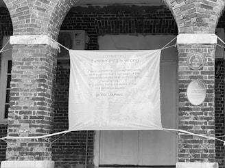 A black and white image showing an architectural setting with two brick arch columns supporting the structure. A large banner is hung between the columns, displaying a quote about the significance of art in society, credited to George Lamming. Plaques on the wall indicate the building's historic interest.