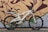 A white electric bicycle is parked against a brick wall with graffiti art. The bike features a sturdy frame, large black wheels, and a comfortable seat. The artwork on the wall is abstract with bold lines and shades of brown, green, and purple.