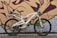 A white electric bicycle is parked against a brick wall with graffiti art. The bike features a sturdy frame, large black wheels, and a comfortable seat. The artwork on the wall is abstract with bold lines and shades of brown, green, and purple.