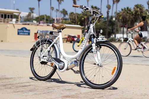 A white electric bicycle with a brown seat is parked on a sandy path near a beach on a sunny day. In the background, palm trees are visible, along with a building and other cyclists passing by.