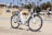A white electric bicycle with a brown seat is parked on a sandy path near a beach on a sunny day. In the background, palm trees are visible, along with a building and other cyclists passing by.
