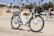 A white electric bicycle with a brown seat is parked on a sandy path near a beach on a sunny day. In the background, palm trees are visible, along with a building and other cyclists passing by.