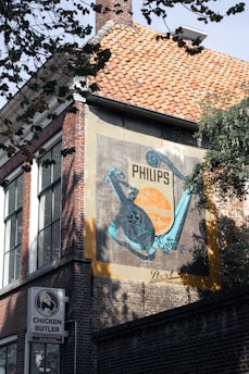 A building with a brick facade prominently features a vintage advertisement for Philips, painted on a large panel. The ad includes a teal-colored graphic design with a musical instrument motif against an orange and black background. Below the ad, a sign for a business called 'Chicken Butler' is visible. Trees partially frame the upper part of the image, casting shadows on the building.