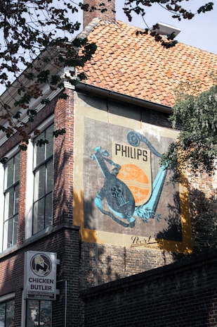 A building with a brick facade prominently features a vintage advertisement for Philips, painted on a large panel. The ad includes a teal-colored graphic design with a musical instrument motif against an orange and black background. Below the ad, a sign for a business called 'Chicken Butler' is visible. Trees partially frame the upper part of the image, casting shadows on the building.