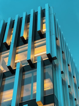 A modern building facade with a grid-like structure. The building features large glass windows with vertical metallic panels. Warm interior lights contrast against the cool blue sky.