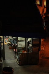 A small, dimly lit store with an awning that obscures most of the upper half. The store's interior is packed with items on shelves and displays. A person is seated inside, legs visible from the doorway. A sign with text is displayed on the glass door, listing an item for sale at 15,000 currency units. The street outside the store is dimly illuminated.