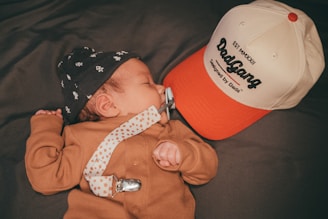 A baby is lying down wearing a patterned hat and a brown onesie. A pacifier is attached to the baby with a clip. Beside the baby is a cap with text on it.