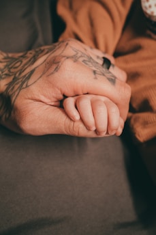 A tattooed adult hand gently holding a small child's hand, conveying a sense of protection and connection. The adult wears a ring, and the child's hand appears delicate against the warmth of the adult's grip.