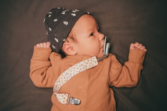 A baby wearing a brown outfit and a patterned hat is lying on a dark surface. The baby has a pacifier attached to a dotted strap clipped to the clothing.
