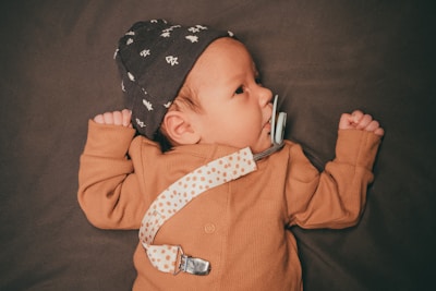 A baby wearing a brown outfit and a patterned hat is lying on a dark surface. The baby has a pacifier attached to a dotted strap clipped to the clothing.