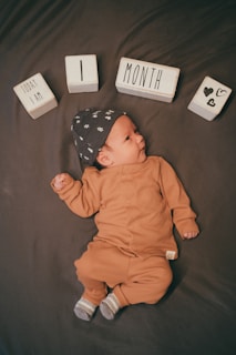 A baby lies on a dark surface, dressed in a brown outfit with a matching hat featuring small designs. Wooden blocks arranged above the baby's head spell out 'Today I Am 1 Month' along with heart symbols.