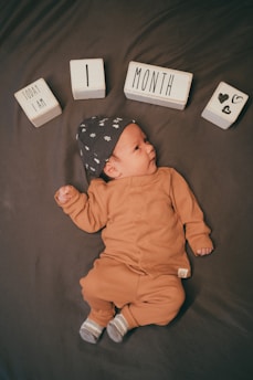 A baby lies on a dark surface, dressed in a brown outfit with a matching hat featuring small designs. Wooden blocks arranged above the baby's head spell out 'Today I Am 1 Month' along with heart symbols.