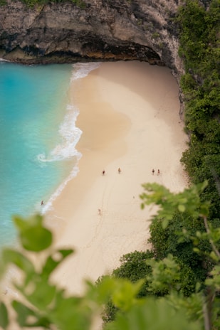 A secluded sandy beach is nestled between tall rocky cliffs and lush greenery. Crystal clear turquoise waves gently lap at the shore. A few people are scattered along the beach, leaving footprints in the sand and enjoying the serene surroundings.