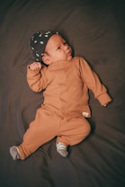 A baby lies on a dark-colored surface, wearing a brown outfit with buttons down the front and a black cap with white patterns. The baby is looking off to the side with a calm expression and is dressed in small striped socks.