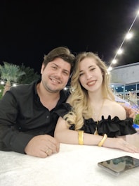 A man and woman are sitting together at a table outdoors at night. They both appear relaxed and happy, with the woman resting her hand on the man's arm. There are string lights above them and a palm tree in the background, suggesting a pleasant outdoor setting.