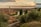 A picturesque view of an Italian city with a river winding through it, surrounded by historic buildings and lush greenery. The setting sun casts a warm glow over the scene, with clouds adding drama to the sky. A stone staircase leads down into the heart of the city.