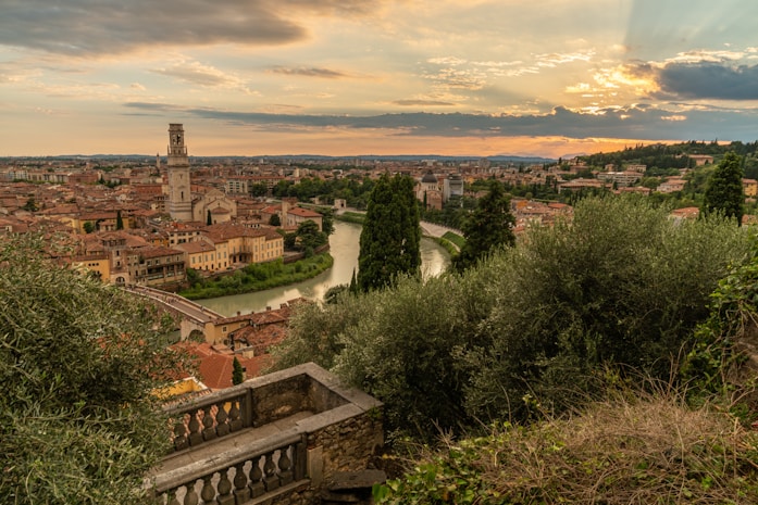 A picturesque view of an Italian city with a river winding through it, surrounded by historic buildings and lush greenery. The setting sun casts a warm glow over the scene, with clouds adding drama to the sky. A stone staircase leads down into the heart of the city.
