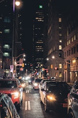 A bustling city street at night with numerous cars lined up, headlights shining brightly. Tall buildings on either side are illuminated, with one displaying a brightly lit 'TD' sign at the top. Streetlights and traffic signals contribute to the urban atmosphere.