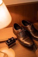 A pair of polished leather dress shoes sit elegantly on a wooden surface next to a small black box containing cufflinks. A warm light from a nearby lamp casts a soft glow over the scene, creating shadows and reflections on the surface.