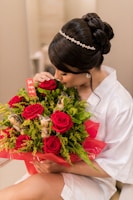 A person with elegant hair adorned with a jeweled band cradles a bouquet of vibrant red roses mixed with lush green foliage and gold-wrapped chocolates. They are dressed in a white garment and are gently smelling the flowers. The bouquet is wrapped in red cellophane and features a small red card.