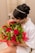 A person with elegant hair adorned with a jeweled band cradles a bouquet of vibrant red roses mixed with lush green foliage and gold-wrapped chocolates. They are dressed in a white garment and are gently smelling the flowers. The bouquet is wrapped in red cellophane and features a small red card.