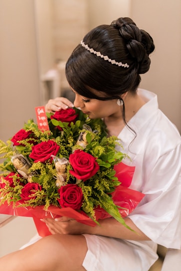 A person with elegant hair adorned with a jeweled band cradles a bouquet of vibrant red roses mixed with lush green foliage and gold-wrapped chocolates. They are dressed in a white garment and are gently smelling the flowers. The bouquet is wrapped in red cellophane and features a small red card.