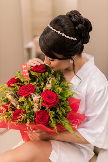 A person with elegant hair adorned with a jeweled band cradles a bouquet of vibrant red roses mixed with lush green foliage and gold-wrapped chocolates. They are dressed in a white garment and are gently smelling the flowers. The bouquet is wrapped in red cellophane and features a small red card.