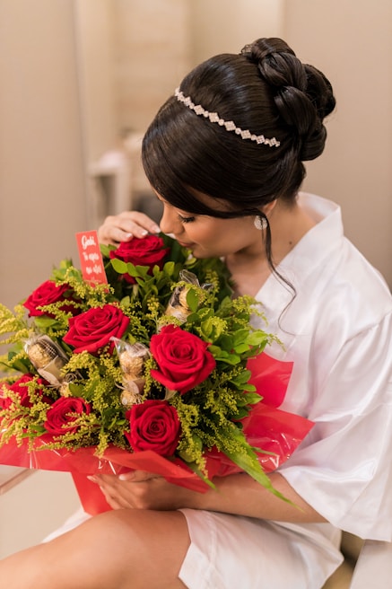 A person with elegant hair adorned with a jeweled band cradles a bouquet of vibrant red roses mixed with lush green foliage and gold-wrapped chocolates. They are dressed in a white garment and are gently smelling the flowers. The bouquet is wrapped in red cellophane and features a small red card.
