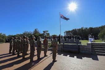A group of soldiers in uniform stands in a formation on a brick pathway under a bright blue sky. Next to them is a flagpole with a flag waving in the wind. In the background, there is a gathering of people, possibly attending a ceremony, on a raised grassy area surrounded by trees. The sun is shining brightly overhead.