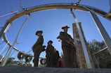 A group of people dressed in military uniforms stand solemnly inside a circular metal structure, with wreaths hanging in the background. The clear blue sky forms a serene backdrop, and a stone pillar is visible in the scene.
