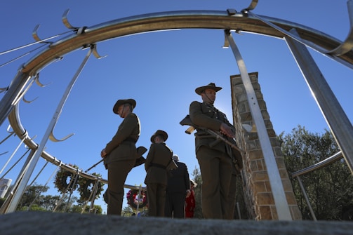 A group of people dressed in military uniforms stand solemnly inside a circular metal structure, with wreaths hanging in the background. The clear blue sky forms a serene backdrop, and a stone pillar is visible in the scene.