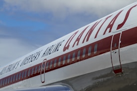 Close-up view of an airplane fuselage with the Qantas logo prominently displayed in large red letters. The aircraft door and passenger windows are visible, with reflections on the shiny metallic surface. The background includes a partly cloudy sky.