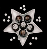 A white, star-shaped dish holds six chocolate truffles, each topped with white icing and decorative sprinkles. Around the dish, white round candies are scattered on a black background, creating a visually striking contrast.