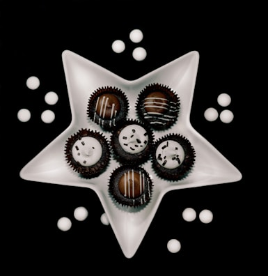 A white, star-shaped dish holds six chocolate truffles, each topped with white icing and decorative sprinkles. Around the dish, white round candies are scattered on a black background, creating a visually striking contrast.