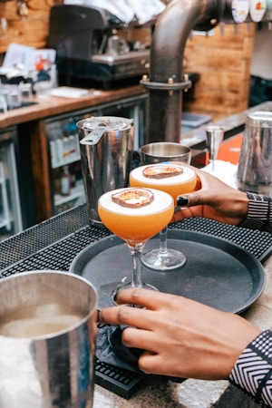 A person is placing a tray with two cocktails on a bar counter. The cocktails are garnished with a slice of citrus fruit on top. The setting is a rustic bar with a wooden counter and metal appliances in the background.