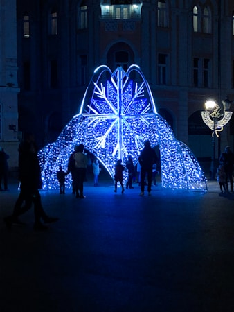 A decorative installation shaped like a snowflake illuminated with blue and white lights stands in an urban setting during the night. Several people, including children, are gathered around the installation, creating a sense of community and exploration.