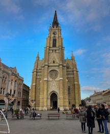 A large, ornate Gothic-style church with a tall spire and clock is centrally situated in a public square. The facade is detailed with intricate stonework, including arches and a large circular window. Surrounding the church, people are engaged in various activities such as walking and sitting on benches. The sky is clear and blue, contrasting with the beige tones of the building and the surrounding architecture.