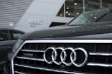 The front grille of an Audi car is prominently displayed, showcasing the company's four-ring logo. The background features the exterior of an Audi dealership, with sleek modern architecture and glass panels.