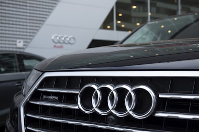 The front grille of an Audi car is prominently displayed, showcasing the company's four-ring logo. The background features the exterior of an Audi dealership, with sleek modern architecture and glass panels.
