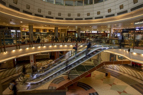 An indoor scene of a shopping mall showcasing multiple levels connected by escalators. The upper level features a food court with various eateries, visible storefronts, and several people seated at tables. The lower area has a patterned tile floor with people in motion, creating a dynamic blur effect, especially on the escalators.