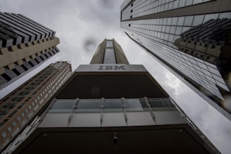 A dramatic perspective of tall, modern skyscrapers converging towards the cloudy sky. The dark tones and reflections on the building surfaces highlight the height and architectural style. The prominent branding of IBM is visible on one of the buildings, creating a strong corporate presence.