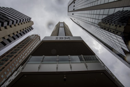 A dramatic perspective of tall, modern skyscrapers converging towards the cloudy sky. The dark tones and reflections on the building surfaces highlight the height and architectural style. The prominent branding of IBM is visible on one of the buildings, creating a strong corporate presence.