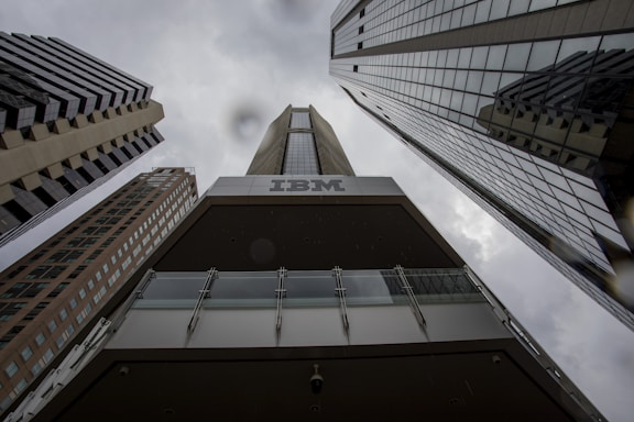 A dramatic perspective of tall, modern skyscrapers converging towards the cloudy sky. The dark tones and reflections on the building surfaces highlight the height and architectural style. The prominent branding of IBM is visible on one of the buildings, creating a strong corporate presence.
