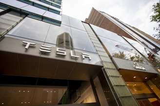A modern building with reflective glass windows and clear signage displaying the name 'TESLA' prominently. The structure has sleek architectural lines and adjacent signage for 'OMEGA'. The background includes other tall buildings and some trees, hinting at an urban setting.