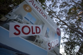 A real estate auction sign displaying the word 'SOLD' in bold red letters. The sign includes images of a home's interior featuring a living room with modern furniture, and it is set against the backdrop of trees and sky.