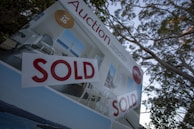 A real estate auction sign displaying the word 'SOLD' in bold red letters. The sign includes images of a home's interior featuring a living room with modern furniture, and it is set against the backdrop of trees and sky.