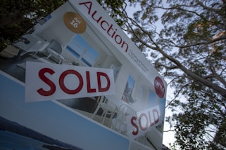 A real estate auction sign displaying the word 'SOLD' in bold red letters. The sign includes images of a home's interior featuring a living room with modern furniture, and it is set against the backdrop of trees and sky.