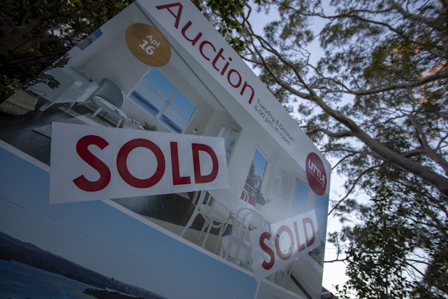 A real estate auction sign displaying the word 'SOLD' in bold red letters. The sign includes images of a home's interior featuring a living room with modern furniture, and it is set against the backdrop of trees and sky.