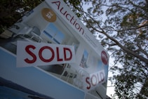 A real estate auction sign displaying the word 'SOLD' in bold red letters. The sign includes images of a home's interior featuring a living room with modern furniture, and it is set against the backdrop of trees and sky.