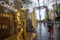 A golden astronaut statue is prominently displayed in a storefront window. The background features a busy city street with people walking by and multiple Omega brand signs visible.